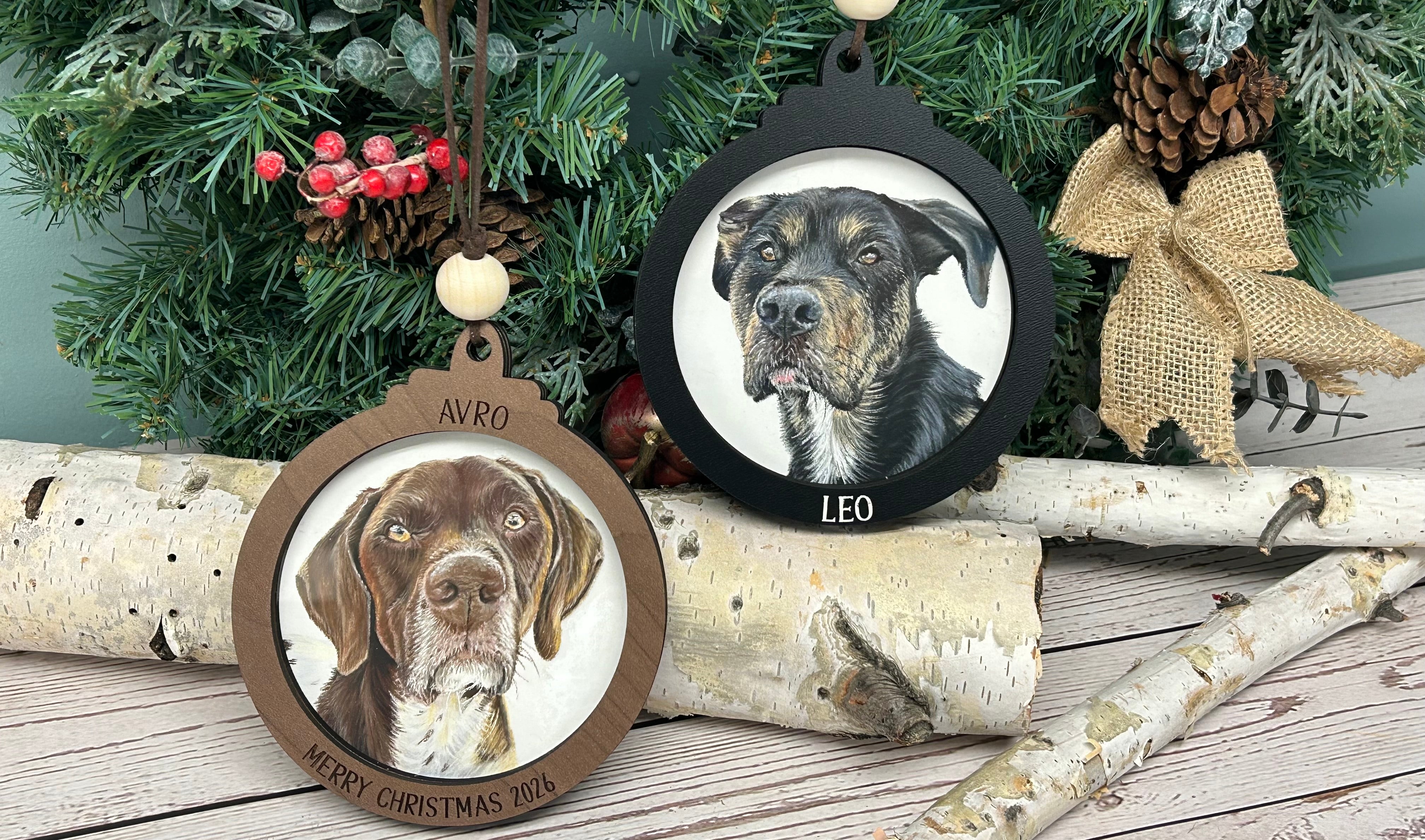 Two round Christmas ornaments featuring custom dog portraits, displayed on birch logs in front of a decorated Christmas tree. One ornament has a light brown wood frame and is labeled 'AVRO', while the other has a black frame and is labeled 'LEO'.