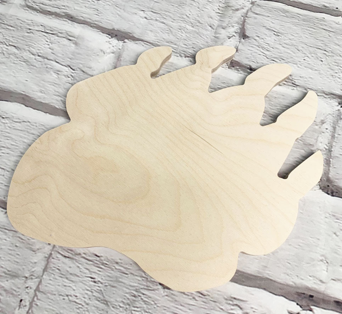 An unfinished light wood bear paw print plaque or cutout with visible natural wood grain, displayed on a white painted brick background.